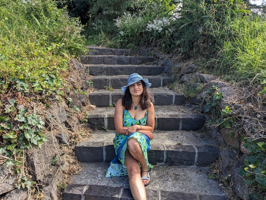Shanti, a young woman with medium length quite dark hair sits on some steps in the sunshine wearing a green slip dress and a slightly wonky homemade indigo hat